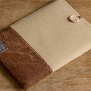 K&ouml;nnte beinhalten: Eine beige und braune Laptop-H&uuml;lle aus Canvas mit einem Knopfverschluss. Die H&uuml;lle hat ein Etikett mit der Aufschrift "POCHETTE GAR&Ccedil;ONNETTE" mit dem Text "Classic & Handmade" und "VIBES" darunter.