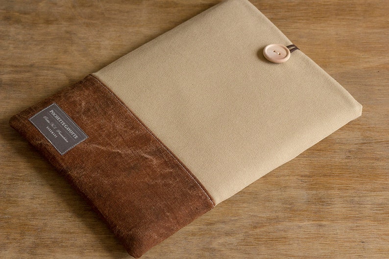 K&ouml;nnte beinhalten: Eine beige und braune Laptop-H&uuml;lle aus Canvas mit einem Knopfverschluss. Die H&uuml;lle hat ein Etikett mit der Aufschrift "POCHETTE GAR&Ccedil;ONNETTE" mit dem Text "Classic & Handmade" und "VIBES" darunter.