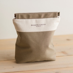 May include: A canvas bag in shades of olive green and cream, with the text "POCHETTE GAVOTTE" in a dark color. The bag has a rectangular shape and a wooden clasp at the top. The bag is sitting on a wooden surface.