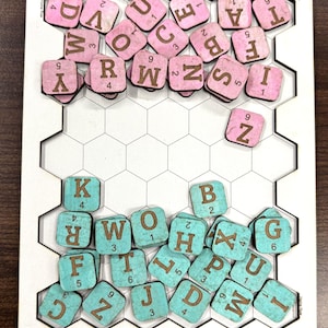 May include: A white rectangular board with a hexagonal grid and the text HomeWords printed on the top and bottom. The board contains pink and turquoise square letter tiles, each featuring a brown letter and a small numerical value in the corner.