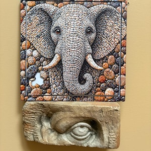 May include: An artistic depiction of an elephant's face, created from small, textured stones in grey, brown, and orange hues. Beneath, a carved stone eye provides a contrasting element. The artwork is displayed on a light-coloured wall.