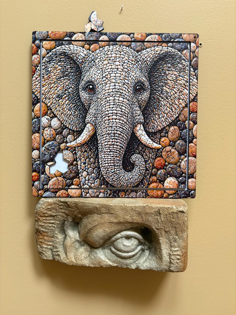 May include: An artistic depiction of an elephant's face, created from small, textured stones in grey, brown, and orange hues. Beneath, a carved stone eye provides a contrasting element. The artwork is displayed on a light-coloured wall.