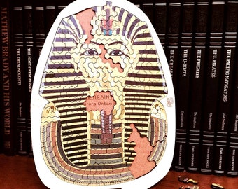 May include: A wooden jigsaw puzzle depicting an Egyptian pharaoh's mask. The puzzle is assembled and features a detailed design with gold and black stripes. The puzzle is on a wooden table with some puzzle pieces scattered around it.