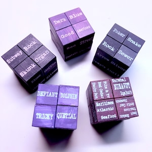 May include: Five wooden cubes with various words printed on each face. The cubes are painted in shades of purple and dark brown. The words include "Dark Blue", "Good American", "Subway Rock", "Defiant Dolphin", and "Narwhal Nunavut".