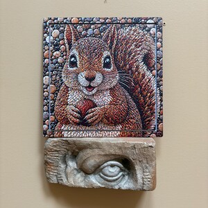 May include: A detailed illustration of a squirrel holding a nut, set against a pebble-textured border. The artwork is mounted above a carved stone eye sculpture, creating a unique contrast in textures and styles.