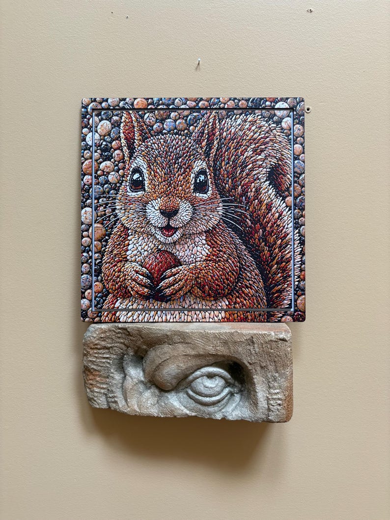 May include: A detailed illustration of a squirrel holding a nut, set against a pebble-textured border. The artwork is mounted above a carved stone eye sculpture, creating a unique contrast in textures and styles.