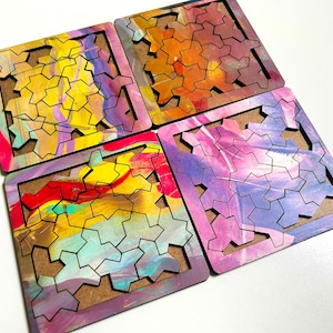 May include: Four square wooden puzzle tiles with a geometric pattern cut out. Each tile has a different abstract design with colorful paint splatters in shades of yellow, red, blue, purple, and orange.