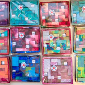 May include: A collection of twelve square wooden perpetual calendar puzzles, each featuring a unique colourful abstract pattern. Each puzzle contains movable blocks, a circular logo sticker, and white squares printed with Thu, Dec, and 25.