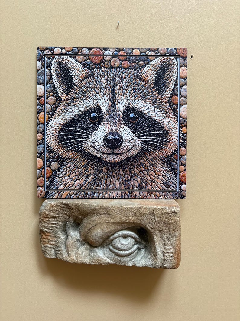 May include: A mosaic artwork depicting a smiling raccoon, framed with stones. The raccoon's fur is a mix of brown, grey, and white. Beneath it, a carved stone eye sculpture is displayed.