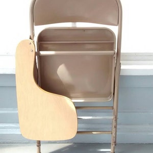 May include: A beige metal folding chair with a wooden writing surface attached to the side. The chair is folded and standing on a white floor.