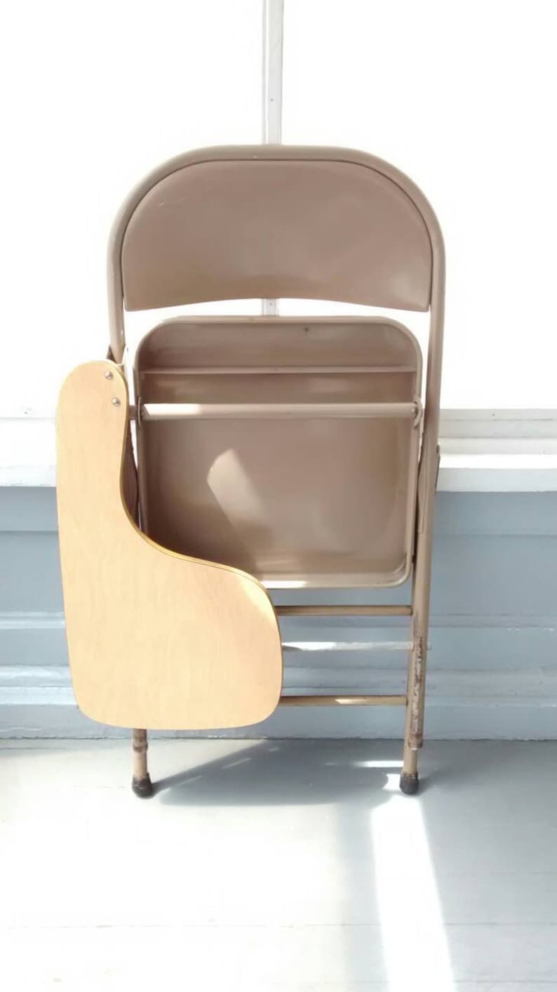 May include: A beige metal folding chair with a wooden writing surface attached to the side. The chair is folded and standing on a white floor.