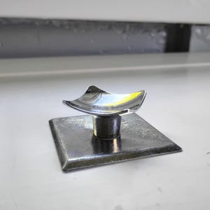May include: A metallic object featuring a flat square base and a central cylindrical post topped with a curved, concave square platform. The piece has a polished, reflective silver-toned finish throughout, with a smooth, uniform metallic texture.
