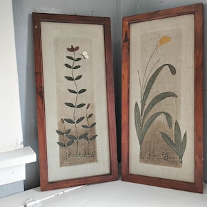 May include: Two framed botanical art pieces. One features a tall plant with dark leaves and a butterfly, the other a yellow flower with green leaves. Both are in rectangular, dark wood frames, leaning against a wall.