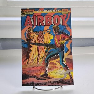 May include: An Eclipse Comics Airboy comic book, number 26, featuring a bi-weekly cover illustration. The artwork depicts a character in a red and blue suit holding a rifle, standing near a cloaked figure and another person in a desert landscape.