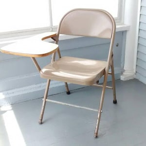 May include: A beige metal folding chair with a white, curved writing surface attached to the right side. The chair is on a light gray floor.