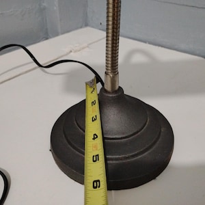 May include: A black desk lamp with a flexible, silver-colored neck and a round, tiered base. The base is approximately 5 inches in diameter. A yellow measuring tape is placed next to the base, and a black power cord is visible.