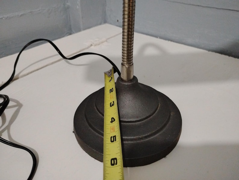 May include: A black desk lamp with a flexible, silver-colored neck and a round, tiered base. The base is approximately 5 inches in diameter. A yellow measuring tape is placed next to the base, and a black power cord is visible.