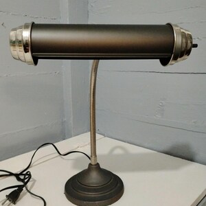 May include: A vintage-style desk lamp with a dark bronze finish. The lamp features a cylindrical shade with silver-colored accents on each end, a flexible gooseneck arm, and a round base. The lamp is plugged into a wall outlet.