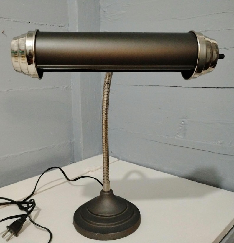 May include: A vintage-style desk lamp with a dark bronze finish. The lamp features a cylindrical shade with silver-colored accents on each end, a flexible gooseneck arm, and a round base. The lamp is plugged into a wall outlet.
