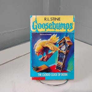 May include: A paperback book titled "Goosebumps: The Cuckoo Clock of Doom" by R.L. Stine. The cover features a yellow and blue design with a bird and a grandfather clock. The book is displayed on a metal stand.