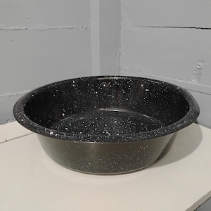 May include: A black enamelware basin with white speckles. The round, open-top bowl has a wide rim and a deep interior. The speckled pattern gives the basin a vintage, rustic aesthetic. Ideal for kitchen or camping use.