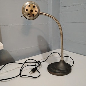 May include: A vintage desk lamp with a flexible, silver-toned neck and a round, bronze-colored base. The lamp head is a metallic cylinder with multiple holes. A black power cord and plug are visible.