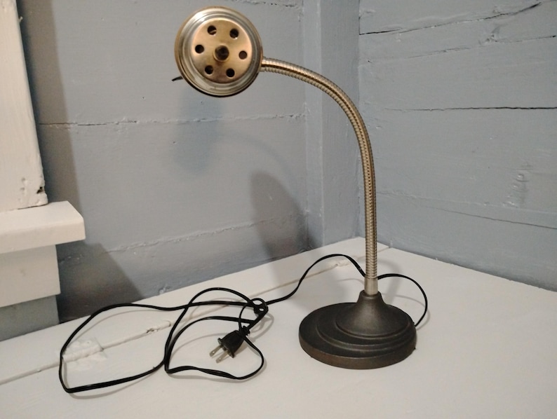 May include: A vintage desk lamp with a flexible, silver-toned neck and a round, bronze-colored base. The lamp head is a metallic cylinder with multiple holes. A black power cord and plug are visible.