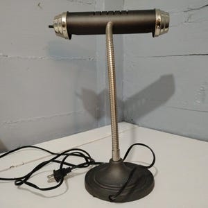 May include: A black desk lamp with a cylindrical shade and a flexible, silver-colored neck. The lamp has a round base and silver accents on the shade. A black power cord extends from the base.