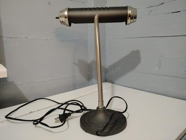 May include: A black desk lamp with a cylindrical shade and a flexible, silver-colored neck. The lamp has a round base and silver accents on the shade. A black power cord extends from the base.