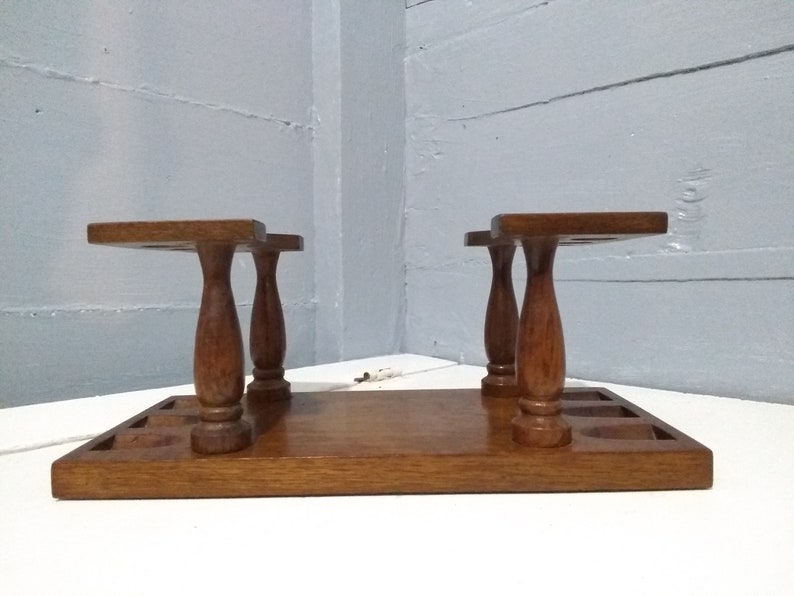 May include: A wooden pipe rack with six slots for pipes. The rack has a brown finish and is made of solid wood. It has a rectangular base and two sets of three turned legs.