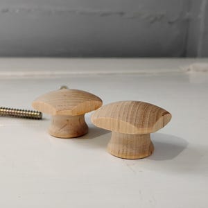 May include: Two unfinished wooden mushroom-shaped cabinet knobs with a screw. The knobs are light brown and have a rounded top and a cylindrical base. The screw is gold-colored.