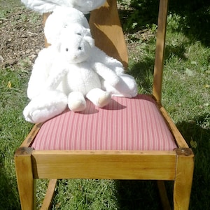 Puede incluir: Un conejito de peluche blanco se sienta en una mecedora de madera con un coj&iacute;n a rayas rojas y blancas. La silla est&aacute; al aire libre sobre una superficie cubierta de hierba.