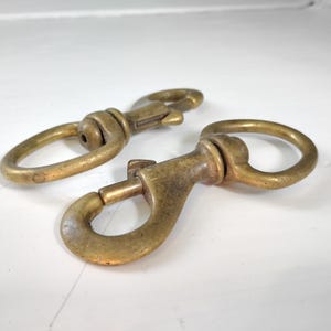 May include: Two antique brass-colored snap hooks. Each hook has a swivel eye and a spring-loaded clasp. The metal has a slightly aged appearance. These hooks are suitable for various applications, such as keychains, pet leashes, or bag straps.