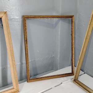 May include: Three antique picture frames. The central frame is a square shape with a gold finish. The other two frames are rectangular with a light brown finish. The frames are empty, with no artwork or glass.