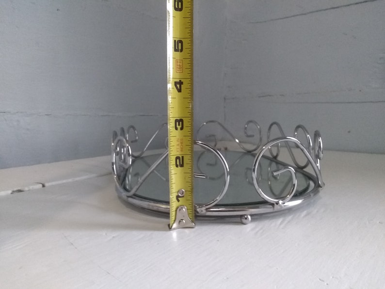 May include: A silver mirrored tray with a decorative scroll design around the edge. The tray is approximately 3 inches tall.