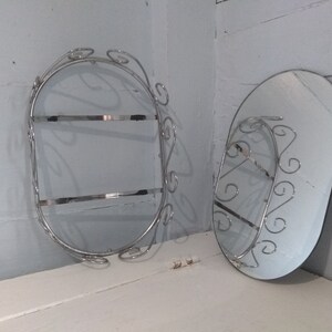 May include: Two oval-shaped wall mounted mirrors with decorative metal frames. The frames are made of silver-colored metal and have a swirling design. The mirrors are both about 12 inches in diameter.
