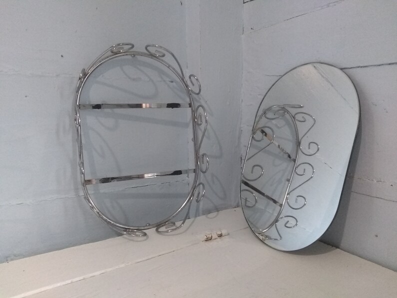 May include: Two oval-shaped wall mounted mirrors with decorative metal frames. The frames are made of silver-colored metal and have a swirling design. The mirrors are both about 12 inches in diameter.