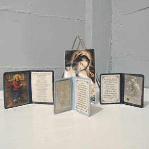 May include: A collection of religious items, including a framed image of Mary and Jesus, and several small, open books with religious imagery and text. The items are displayed on a white surface against a light gray background.