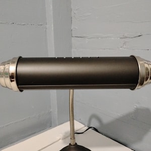 May include: A black desk lamp with a cylindrical shade and silver-colored end caps. The lamp has a flexible, silver-colored gooseneck and a round base. The lamp is on a white surface against a gray wall.
