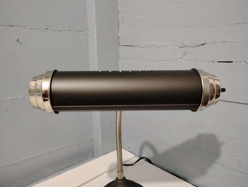 May include: A black desk lamp with a cylindrical shade and silver-colored end caps. The lamp has a flexible, silver-colored gooseneck and a round base. The lamp is on a white surface against a gray wall.