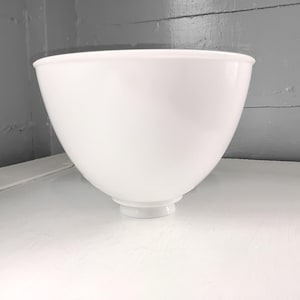 May include: A white, opaque glass bowl with a rounded shape and a flared rim. The bowl has a small, circular base and sits on a white surface. The background is a gray wall and white surface.