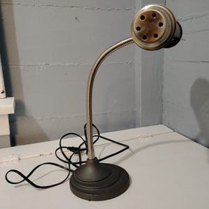 May include: An antique desk lamp with a flexible, silver-colored neck and a round, bronze-toned head. The lamp has a dark gray, circular base and a black power cord. The lamp is positioned on a white surface against a gray wall.