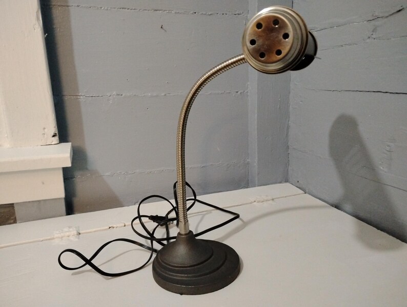 May include: An antique desk lamp with a flexible, silver-colored neck and a round, bronze-toned head. The lamp has a dark gray, circular base and a black power cord. The lamp is positioned on a white surface against a gray wall.