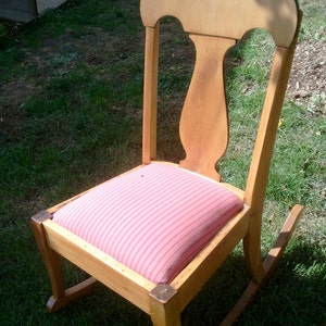 Puede incluir: Una silla mecedora de madera con un coj&iacute;n a rayas rosa y blanco. La silla tiene un respaldo alto y un asiento curvo.
