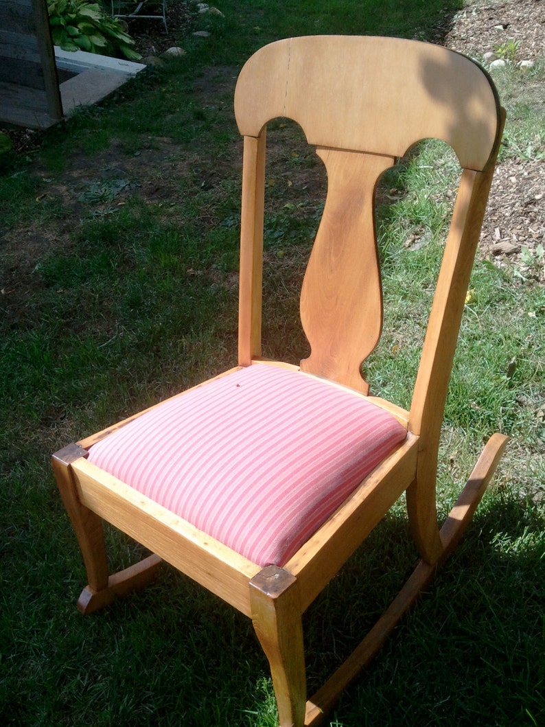Puede incluir: Una silla mecedora de madera con un coj&iacute;n a rayas rosa y blanco. La silla tiene un respaldo alto y un asiento curvo.