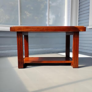 May include: A rectangular, wooden coffee table with a rich, reddish-brown finish. The table features a lower shelf and vertical supports with a grooved design. The table is approximately 48 inches long, 20 inches wide, and 18 inches high.