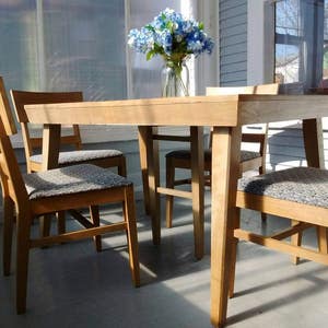 May include: A light-toned wooden dining set consisting of a rectangular table and four chairs. Each chair features a high backrest and a seat upholstered in a grey and white patterned fabric. A clear glass vase with blue and white flowers sits on the table.