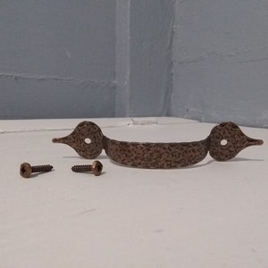 May include: A hammered brass cabinet pull with two screws. The pull has a curved shape with a decorative design and two rounded ends.