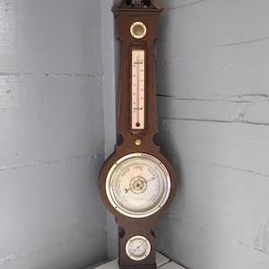 May include: A wall-mounted weather station featuring a dark wood frame with a vertical thermometer, a central circular barometer with a brass-colored bezel and white dial, and a smaller circular hygrometer at the bottom with brass accents.