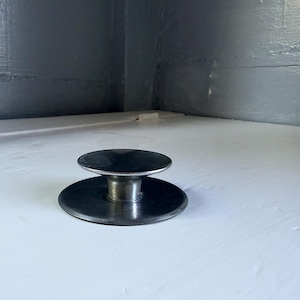 May include: A dark metallic circular knob featuring a flat base and a slightly concave top surface connected by a central cylindrical stem. The object has a smooth, reflective finish and a uniform dark gray color throughout its entire structure.